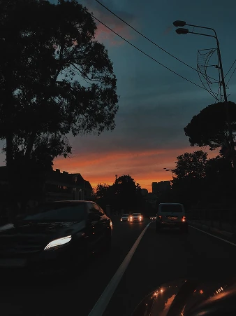Car headlights on a road at dusk, under silhouetted trees and an orange sky