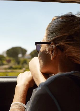 A woman in sunglasses resting her arms on an open car window, looking out at the countryside